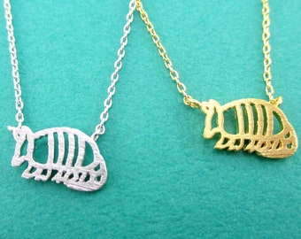 Peut inclure: Deux colliers avec pendentifs en forme de tatou. L'un est argenté, l'autre doré. Les deux colliers ont une chaîne délicate et un pendentif silhouette de tatou détaillé. Le fond est de couleur turquoise uni.