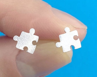 May include: Two small, silver-colored puzzle piece stud earrings. The earrings are in the shape of a jigsaw puzzle piece, with a brushed metal finish. The earrings are displayed against a blue background.