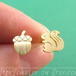 May include: Gold-colored stud earrings featuring an acorn and a squirrel. The acorn earring has a textured cap and a smooth, vertical-lined body. The squirrel earring is a silhouette with a detailed tail. The Etsy shop name, "Atti Jewelry on Etsy.com", is visible.