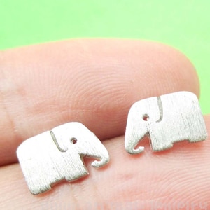 May include: Two small, silver-colored elephant-shaped charms. Each charm has a textured surface and a simple, stylized design with a trunk, eye, and ear detail. The charms are shown against a green background.