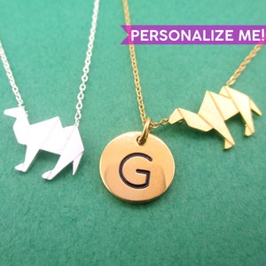 Peut inclure: Deux colliers avec des pendentifs en forme de chameau. L'un est argenté, l'autre doré. Le collier doré a un pendentif rond avec la lettre "G". Le fond est vert uni. Le texte "PERSONNALISEZ-MOI !" est dans une bannière violette.