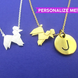 Peut inclure: Trois colliers avec des pendentifs en forme de caniche origami et un pendentif rond avec la lettre "J". Un collier est argenté, les deux autres sont dorés. Les pendentifs sont suspendus à de fines chaînes sur fond bleu.