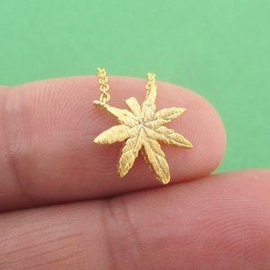 May include: A gold-colored necklace featuring a detailed cannabis leaf pendant. The pendant is suspended from a delicate chain. The necklace is shown against a green background.