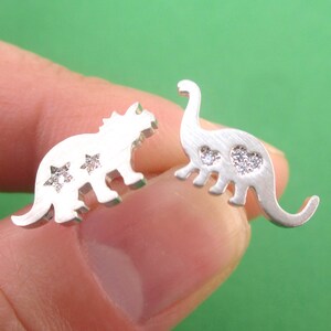 May include: A pair of silver-colored dinosaur stud earrings. One earring is a triceratops shape with star-shaped accents, and the other is a brontosaurus shape with heart-shaped accents. The earrings are held against a green background.