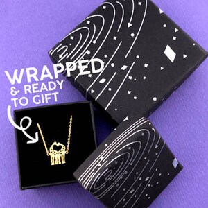May include: A gold-colored necklace with a giraffe pendant is displayed in a black gift box. The box is open, revealing the necklace. The box is on a purple surface, with two black gift boxes with a white galaxy design. The text "Wrapped & Ready to Gift" is visible.