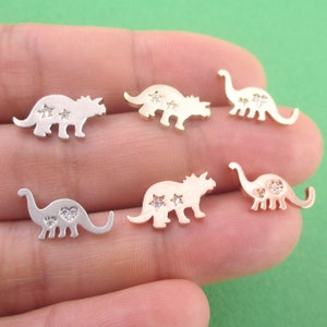 May include: A collection of small dinosaur-shaped charms in silver and gold tones. Some charms feature star or heart-shaped embellishments. The charms are displayed against a green background.