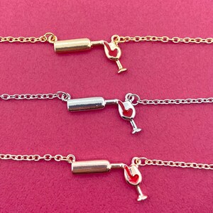 Op de afbeelding: Drie delicate kettingen met een wijnfles- en wijnglasbedel. De bedels zijn goud- en zilverkleurig, met een rood emaille detail in het wijnglas. De kettingen worden getoond op een roze achtergrond.