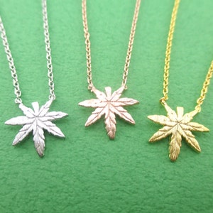 May include: Three necklaces with leaf pendants in silver, rose gold, and gold colors. Each pendant is a detailed cannabis leaf shape, suspended from a delicate chain. The necklaces are displayed against a green background.
