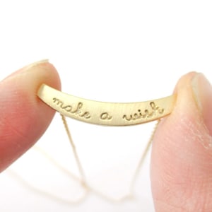 Puede incluir: Un collar dorado con un colgante de barra curvada. El colgante está inscrito con las palabras "make a wish" en una fuente delicada y manuscrita. El collar se sostiene entre dos dedos sobre un fondo blanco.