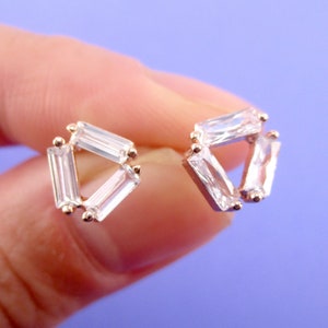 May include: A pair of gold-toned stud earrings, each featuring a triangular arrangement of three clear, rectangular-cut gemstones. The earrings are set against a light purple background.
