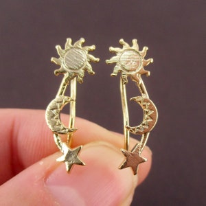 May include: A pair of gold-colored ear climbers featuring celestial designs. Each earring has a sun at the top, a crescent moon in the middle, and a star at the bottom. The earrings are designed to climb up the ear.