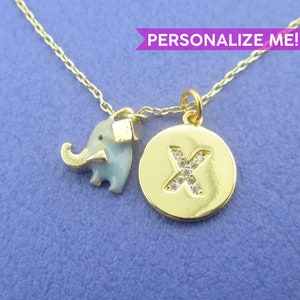 Puede incluir: Collar dorado con dos colgantes: un elefante azul claro y un colgante redondo con la letra "X" incrustada con pequeñas piedras transparentes. El texto "PERSONALIZE ME!" está en una pancarta morada.