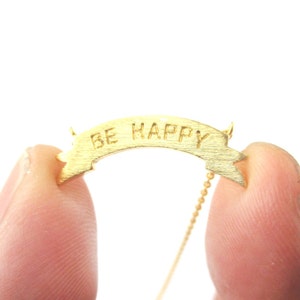 Puede incluir: Un collar dorado con un colgante de banner. El banner dice "BE HAPPY" en letras mayúsculas grabadas. El collar se sostiene entre dos dedos sobre un fondo blanco. La cadena es delicada y dorada.