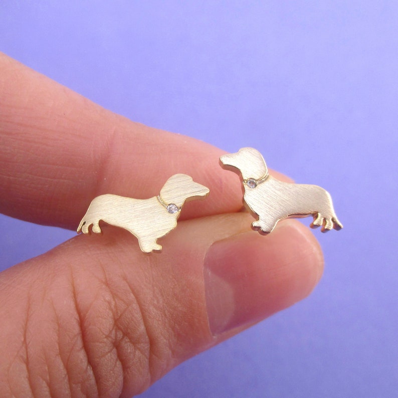 May include: A pair of gold-colored dachshund stud earrings. Each earring features a dog silhouette with a small, clear gem on the collar. The earrings are displayed against a purple background.