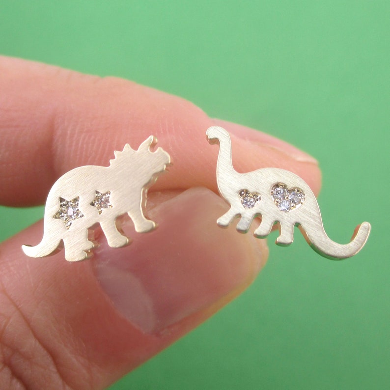 May include: A pair of gold-colored dinosaur stud earrings. One earring is a triceratops shape with two star-shaped crystal accents. The other earring is a long-necked dinosaur shape with two heart-shaped crystal accents.