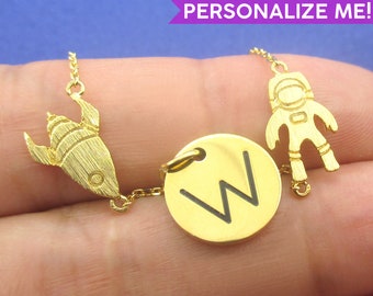 May include: Gold-colored necklace with a rocket, astronaut, and a circular pendant with the letter "W". The necklace is on a chain and the text "PERSONALIZE ME!" is in a purple banner.