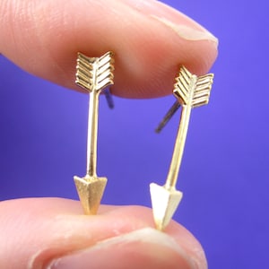 Puede incluir: Un par de pendientes de botón dorados con forma de flecha. Cada pendiente presenta un diseño de flecha detallado con punta y plumas. Los pendientes se sostienen sobre un fondo azul.