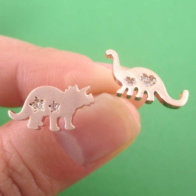 May include: Rose gold dinosaur stud earrings. One earring is a Triceratops with star-shaped cutouts and the other is a Brontosaurus with heart-shaped cutouts. Both earrings have small, clear gemstones.