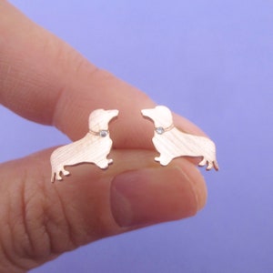 May include: A pair of rose gold dachshund stud earrings. Each earring features a dachshund silhouette with a small, clear gemstone collar. The earrings are held against a purple background.