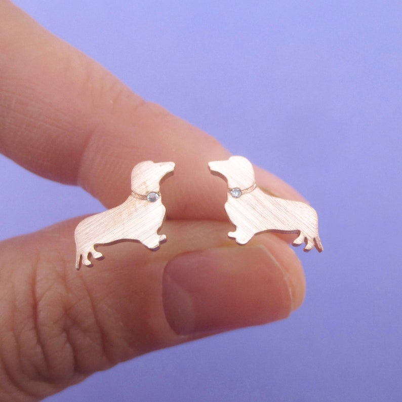 May include: A pair of rose gold dachshund stud earrings. Each earring features a dachshund silhouette with a small, clear gemstone collar. The earrings are held against a purple background.