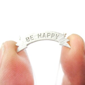 Puede incluir: Un pequeño colgante de banner plateado con las palabras "BE HAPPY" grabadas. El banner se sostiene entre dos dedos sobre un fondo blanco. El colgante tiene un acabado de metal cepillado.