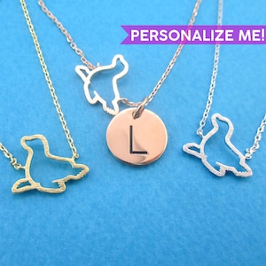 Op de afbeelding: Drie kettingen met zeehondhangerontwerpen. Eén is goudkleurig, één is zilverkleurig en één is roségoudkleurig met een ronde hanger met de letter "L". De tekst "PERSONALIZE ME!" staat in een paarse banner.