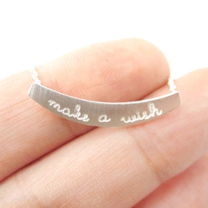 Puede incluir: Un collar plateado con un colgante curvo con la inscripción "make a wish" en escritura cursiva. La joya se muestra sobre un fondo blanco.