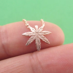 May include: A gold-colored necklace featuring a detailed cannabis leaf pendant. The pendant is intricately designed with visible veins and textures, suspended from a delicate chain. The necklace is shown against a green background.