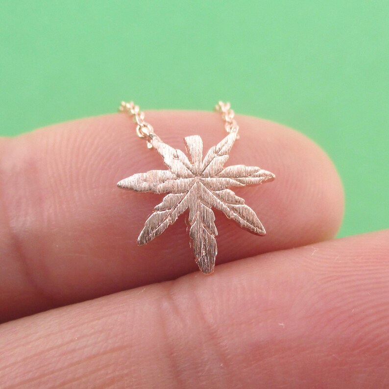 May include: A gold-colored necklace featuring a detailed cannabis leaf pendant. The pendant is intricately designed with visible veins and textures, suspended from a delicate chain. The necklace is shown against a green background.