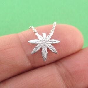 May include: A silver-colored necklace featuring a detailed cannabis leaf pendant. The pendant is intricately designed with textured lines, suspended from a delicate chain. The necklace is displayed against a green background.