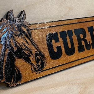 May include: Wooden sign featuring a detailed horse head carving on the left and the name "CURLY" in bold black letters on a rectangular plaque. The sign has a warm, brown finish, with a darker border.
