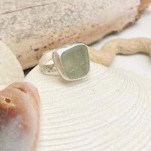 Puede incluir: Un anillo de plata con una piedra cuadrada de color verde mar. El anillo se encuentra sobre una gran concha blanca, con otras conchas y madera flotante en el fondo. La banda del anillo tiene un diseño texturizado.