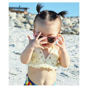 May include: A young child wearing pink heart-shaped sunglasses and a cream-colored crochet halter top. The child is on a sandy beach, with the ocean and rocks in the background. The child's hair is styled in two pigtails.