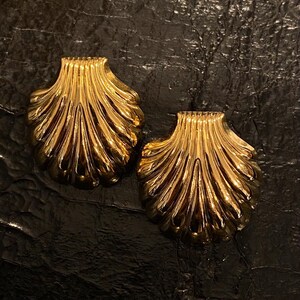 Peut inclure: Une paire de boucles d'oreilles en forme de coquille de couleur or. Les boucles d'oreilles sont en métal et présentent un design détaillé et texturé.
