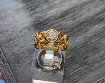 Op de afbeelding: Een goudkleurige ring met een grote, heldere, ronde edelsteen in het midden. De ring heeft een uniek, getextureerd ontwerp op de band. De ring wordt gepresenteerd op een heldere, rechthoekige standaard.