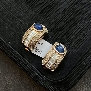 May include: A pair of gold hoop earrings with a blue oval stone and clear crystal accents. The earrings are on a black surface with a white tag that says "S&S 40".