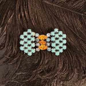 May include: A turquoise and orange rhinestone brooch in the shape of a bow. The brooch is set on a black background.