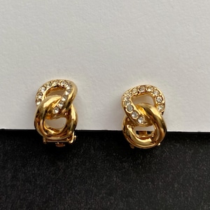 Peut inclure: Une paire de boucles d'oreilles dorées à clip. Chaque boucle d'oreille présente un design à double boucle, dont une incrustée de petits cristaux transparents. Les boucles d'oreilles sont posées sur un fond blanc et noir.