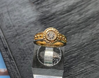 Op de afbeelding: Een goudkleurige ring met een centrale, heldere edelsteen. De ring heeft een gedetailleerd ontwerp rond de edelsteen en langs de band, met kleine, verhoogde cirkelvormige accenten. De ring wordt gepresenteerd op een heldere, rechthoekige standaard.
