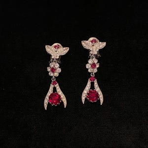 Könnte beinhalten: Ein Paar silberfarbene Ohrhänger mit floralem Design. Jeder Ohrring hat einen großen, runden, roten Edelstein am unteren Ende, mit kleineren roten Edelsteinen und klaren Kristallen. Die Ohrringe haben eine schmetterlingsförmige Oberseite.
