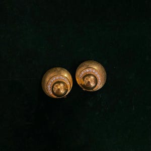 Peut inclure: Une paire de boucles d'oreilles clips en or avec un design rond et des strass transparents.
