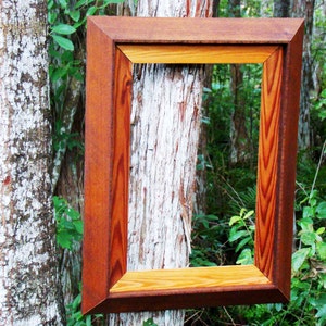 Pu&ograve; includere: Una cornice per foto in legno con una finitura in legno naturale. La cornice &egrave; realizzata con due tipi di legno diversi, uno con una tonalit&agrave; rossiccia pi&ugrave; scura e l'altro con un colore pi&ugrave; chiaro e naturale. La cornice &egrave; appesa a un tronco d'albero in un ambiente boschivo.
