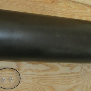 May include: A dark gray metal pipe with a ribbed texture. The pipe is about 12 inches long and has a diameter of about 6 inches. It is lying on a wooden surface.
