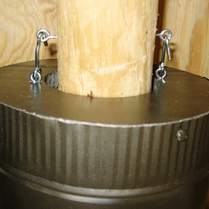 May include: A close-up of a metal chimney pipe with a ribbed surface. The pipe is attached to a wooden post with two metal hooks.