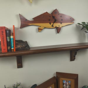 May include: A wooden fish-shaped clock with Roman numerals hangs above a dark brown wooden shelf. The shelf holds books and a small plant in a white pot. The clock is made of two tones of wood, brown and yellow.