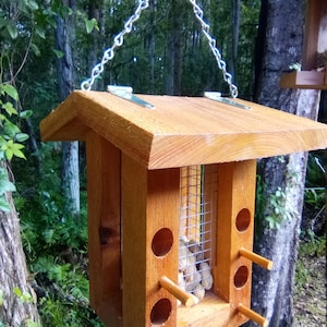 May include: A wooden bird feeder with a wire mesh compartment for peanuts. The feeder has a roof and is hanging from a chain. There are four small wooden perches on the front of the feeder.