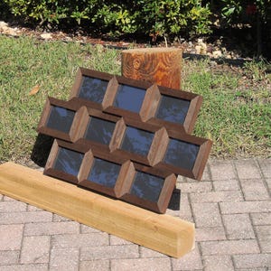 May include: A collection of nine dark brown wooden picture frames with a rectangular design, arranged in a grid. A long, light brown wooden beam rests below the frames. The frames are displayed outdoors on a brick surface.