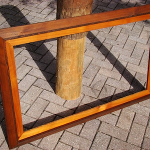 May include: A large, rectangular wooden picture frame with a dark brown outer frame and a lighter brown inner frame. The frame is sitting on a brick patio.
