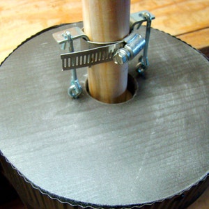 May include: A close-up of a gray, round, corrugated base with a wooden pole secured to it using a metal clamp. The clamp has a series of teeth and a screw.