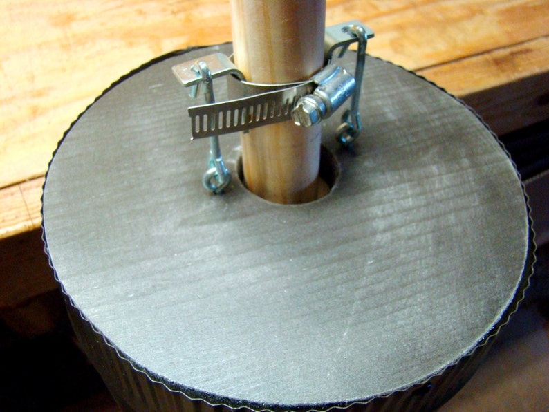 May include: A close-up of a gray, round, corrugated base with a wooden pole secured to it using a metal clamp. The clamp has a series of teeth and a screw.